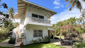 Une maison blanche à deux étages, dotée d’un balcon, entourée d’une végétation tropicale et bordée d’un chemin en pierre.