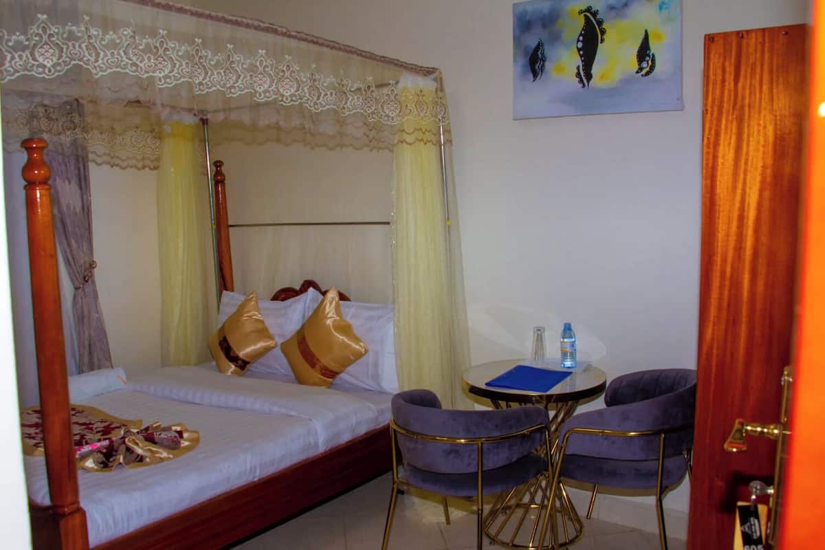 A hotel room with a four-poster bed, two chairs, a small table, and a painting on the wall.