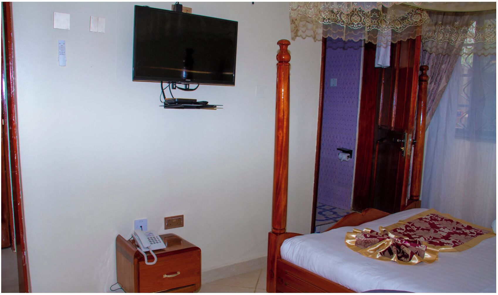 Un dormitorio con cama con dosel, una mesita de noche con teléfono y un televisor montado en la pared.