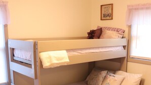 Una cama litera con colchón blanco, almohadas y una manta. Hay una mesita de noche pequeña y blanca con cajones. Un tapete morado está en el suelo.