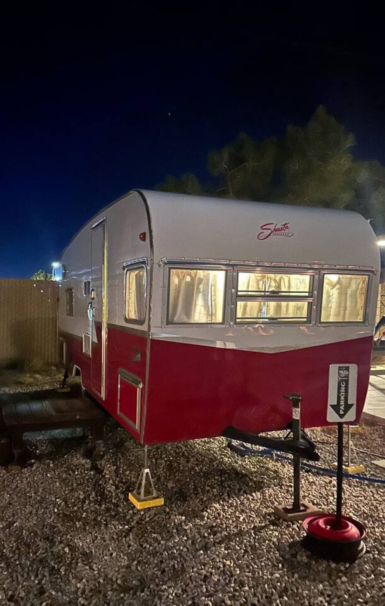 Ein altmodischer Wohnwagen mit roter und weißer Außenfarbe, eine kleine angebaute Hütte und ein Schild, das es als Stellplatz kennzeichnet.
