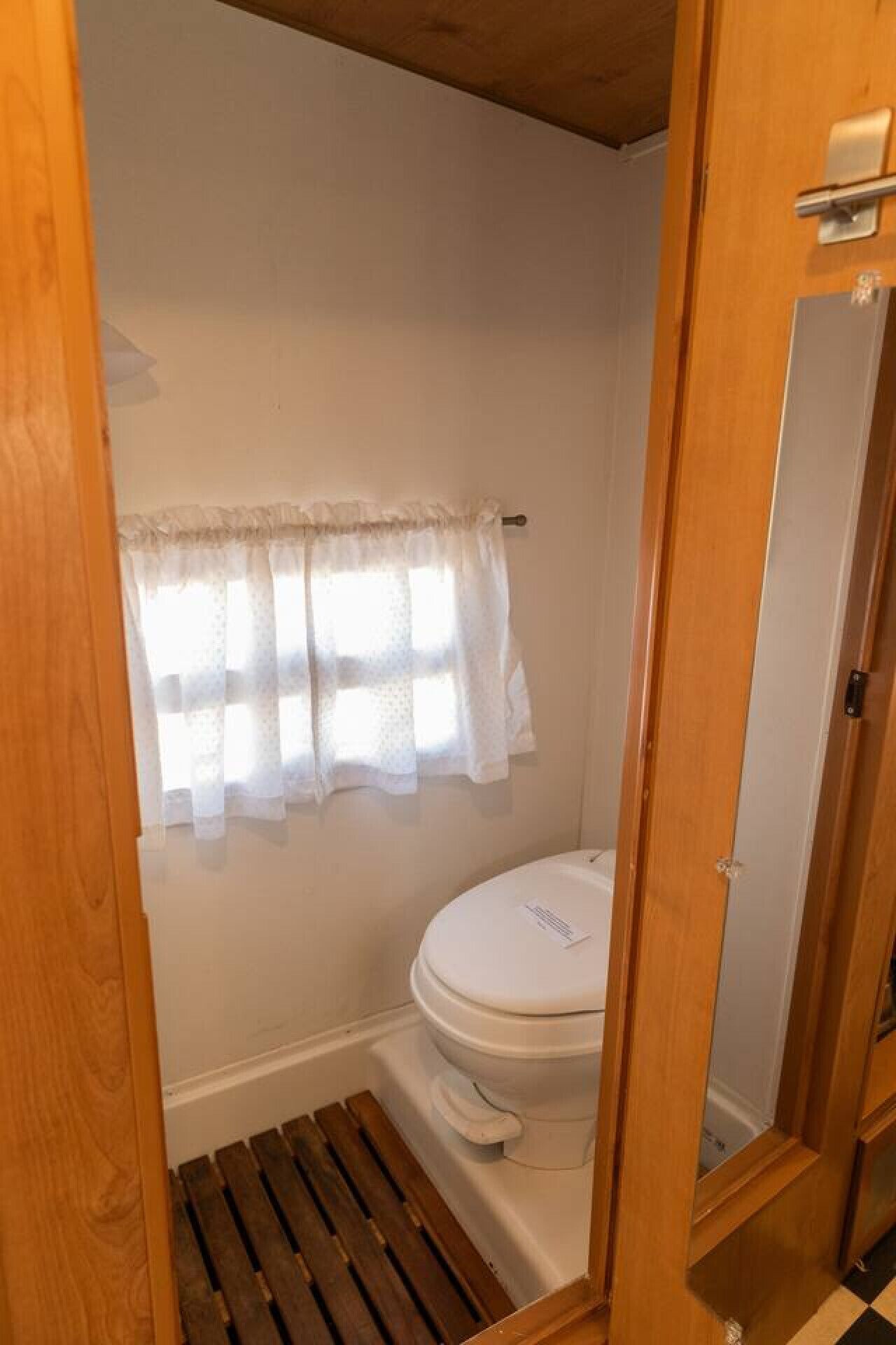 Ein kleines, kompaktes Badezimmer mit Toilette, einer Duschfläche aus Holzlamellen und einem Fenster mit leichtem Vorhang.