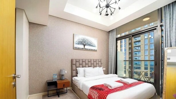 A modern hotel room with a large bed, a nightstand, a painting of a tree, and a view of the city through large windows. - Helnan Royale (Dubai)