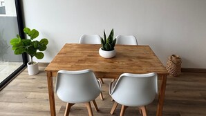 Uma sala de jantar com mesa de madeira, cadeiras brancas, uma planta em vaso e uma cesta de fibra.