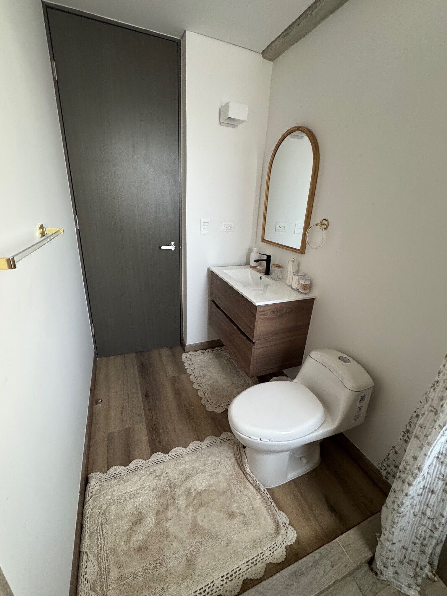 Ein kleines Badezimmer mit Holzboden, Toilette in Weiß, Waschtisch aus Holz, einem Spiegel und einer Tür.
