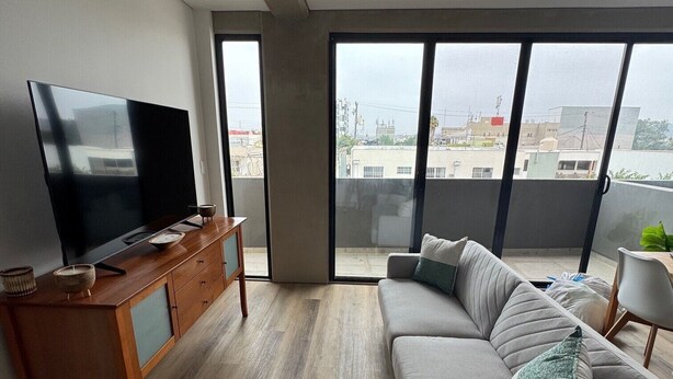 Uma sala de estar moderna com uma TV de tela plana em um armário de madeira, um sofá cinza com almofadas e janelas amplas com vista para a cidade.