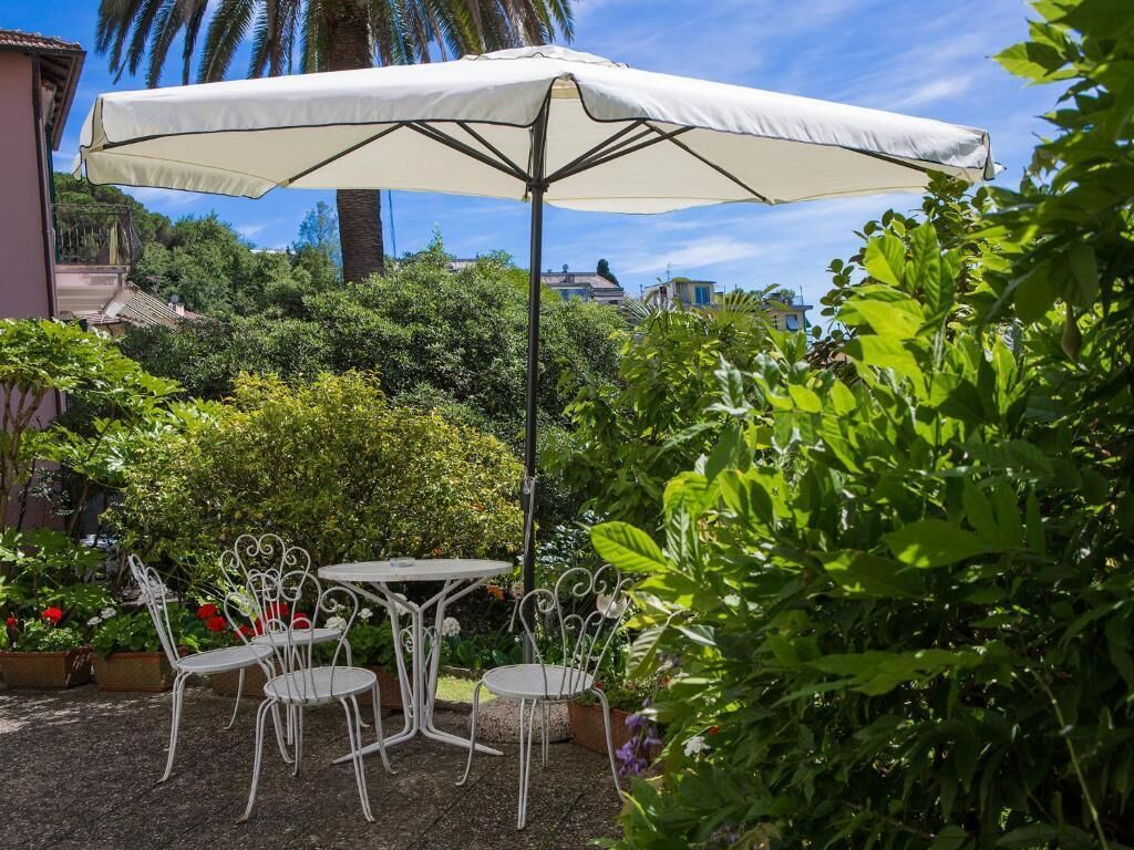 An outdoor patio with a table and chairs under a large umbrella, surrounded by lush greenery and flowering plants.