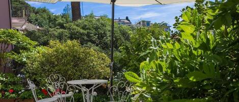 Een terras buiten met een tafel en stoelen onder een grote parasol, omgeven door weelderig groen en bloeiende planten.