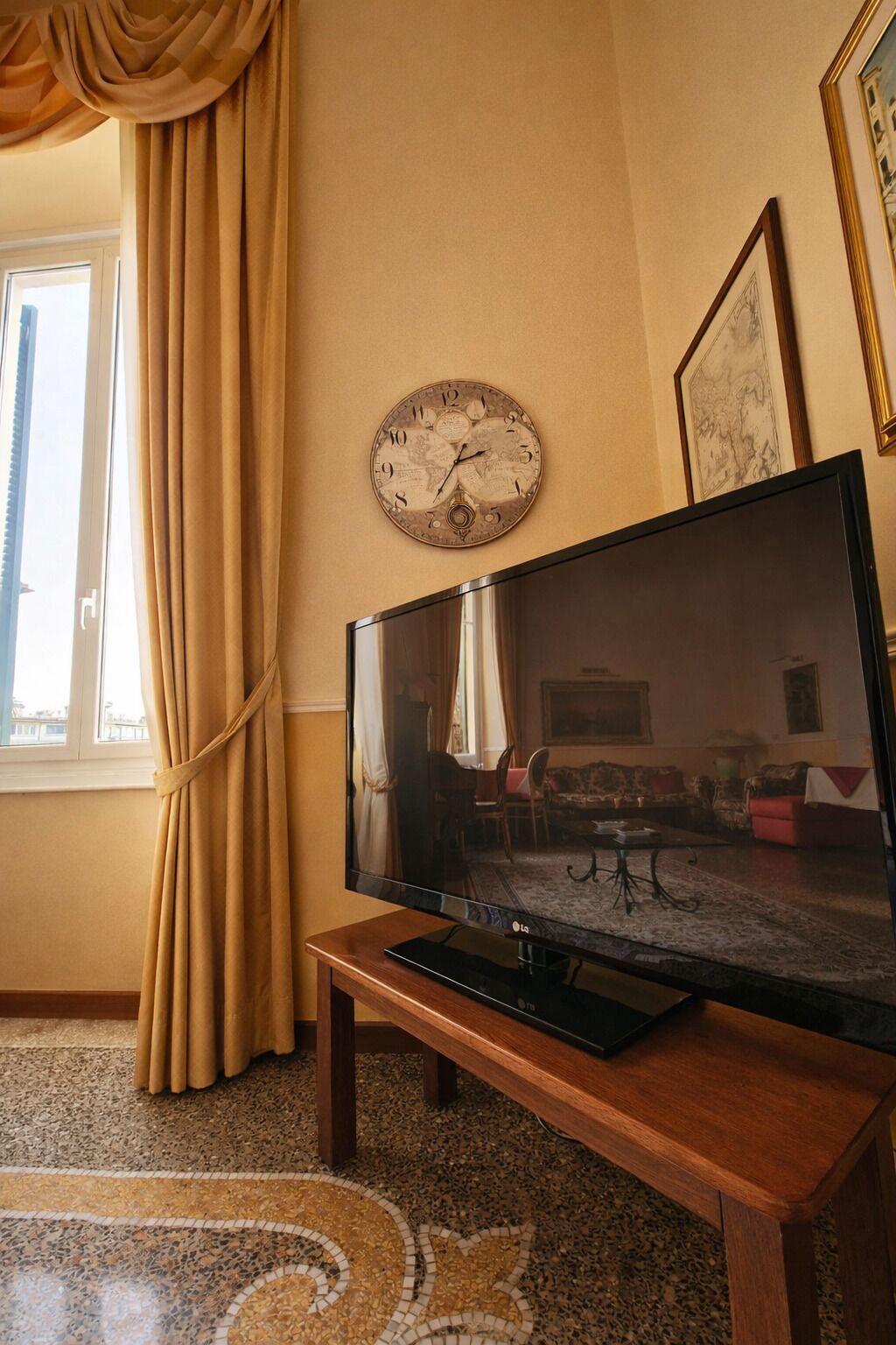 A living room with a flat-screen TV on a wooden stand, a world map clock on the wall, and a large window with curtains.