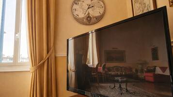 Ein Wohnzimmer mit Flachbildfernseher auf Holzfuß, einer Weltkartenuhr an der Wand und einem großen Fenster mit Vorhängen.