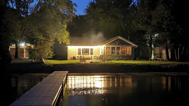 A house with a dock extending into a calm body of water, surrounded by trees. - Peace Cottage on the Water-Rustic Charm & Lakefront Living!! (Faribault)