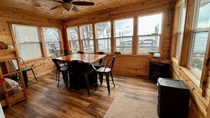A wooden cabin interior with a dining table, chairs, a fireplace, and large windows offering views of the outdoors. - Peace Cottage on the Water-Rustic Charm & Lakefront Living!! (Faribault)