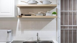 Une cuisine dotée d’un évier à deux bacs, de meubles blancs et d’une étagère où sont rangées des casseroles et une plante.