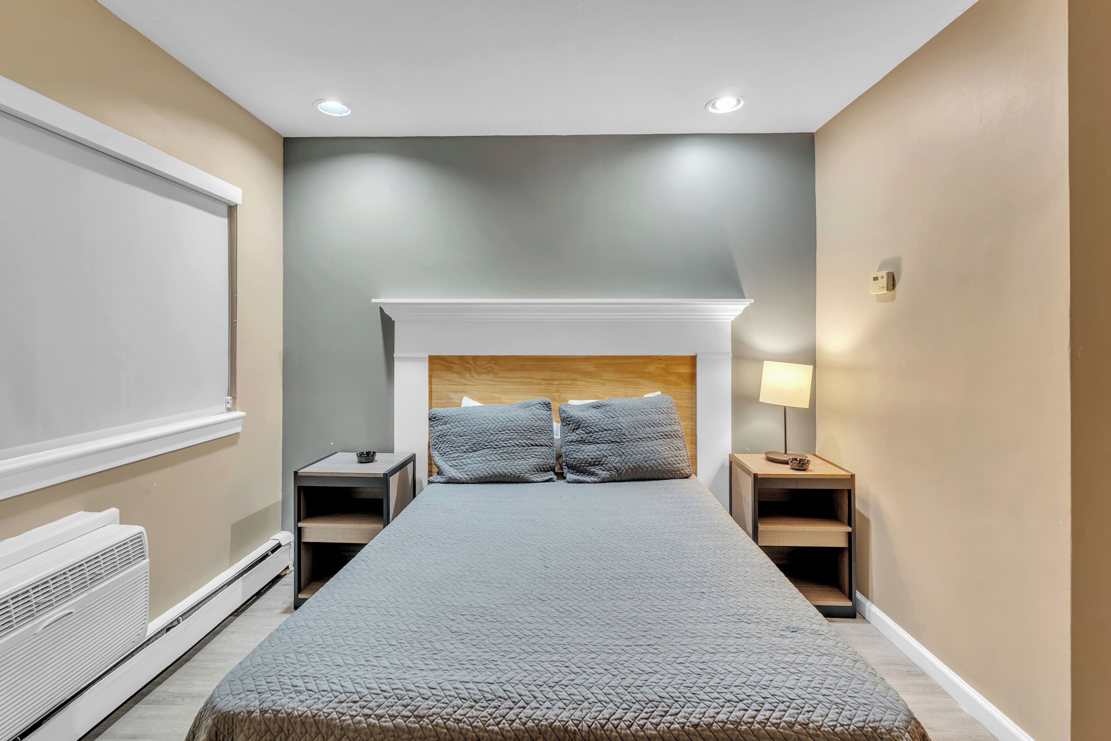 A bedroom with a bed, two bedside tables, a nightstand, a wall-mounted heater, and a ceiling light.