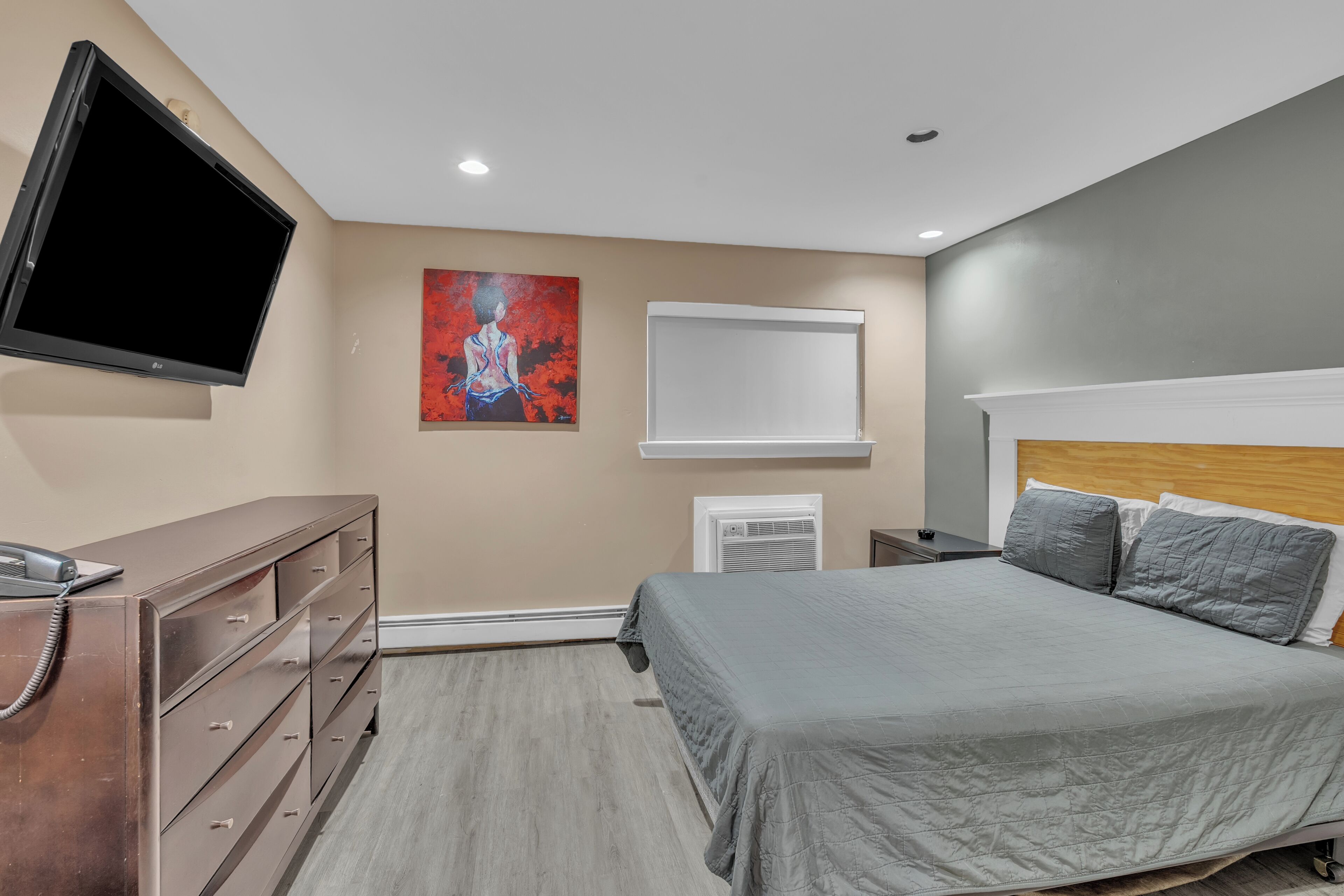 A bedroom with a bed, dresser, TV, artwork, and an air conditioning unit.