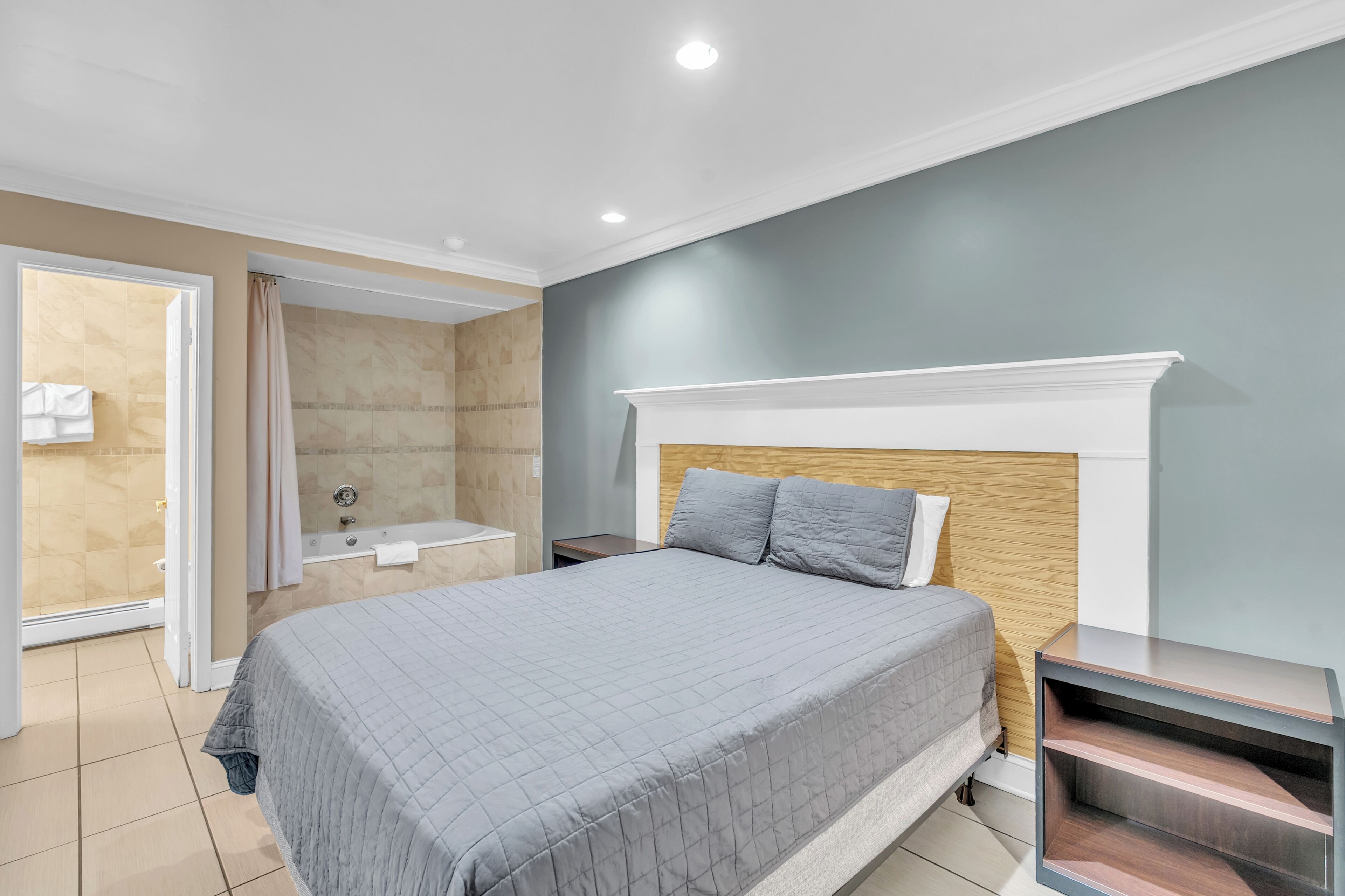 Un dormitorio con cama, mesita de noche, cabecera de madera, estantería empotrada y baño con bañera y ducha.