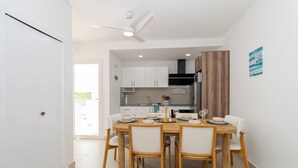 A dining area with a round table and six chairs, a kitchen in the background, and a ceiling fan.