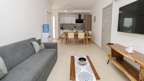 A compact living space with a grey sofa, a dining table with chairs, a kitchen area, and a TV mounted on the wall.