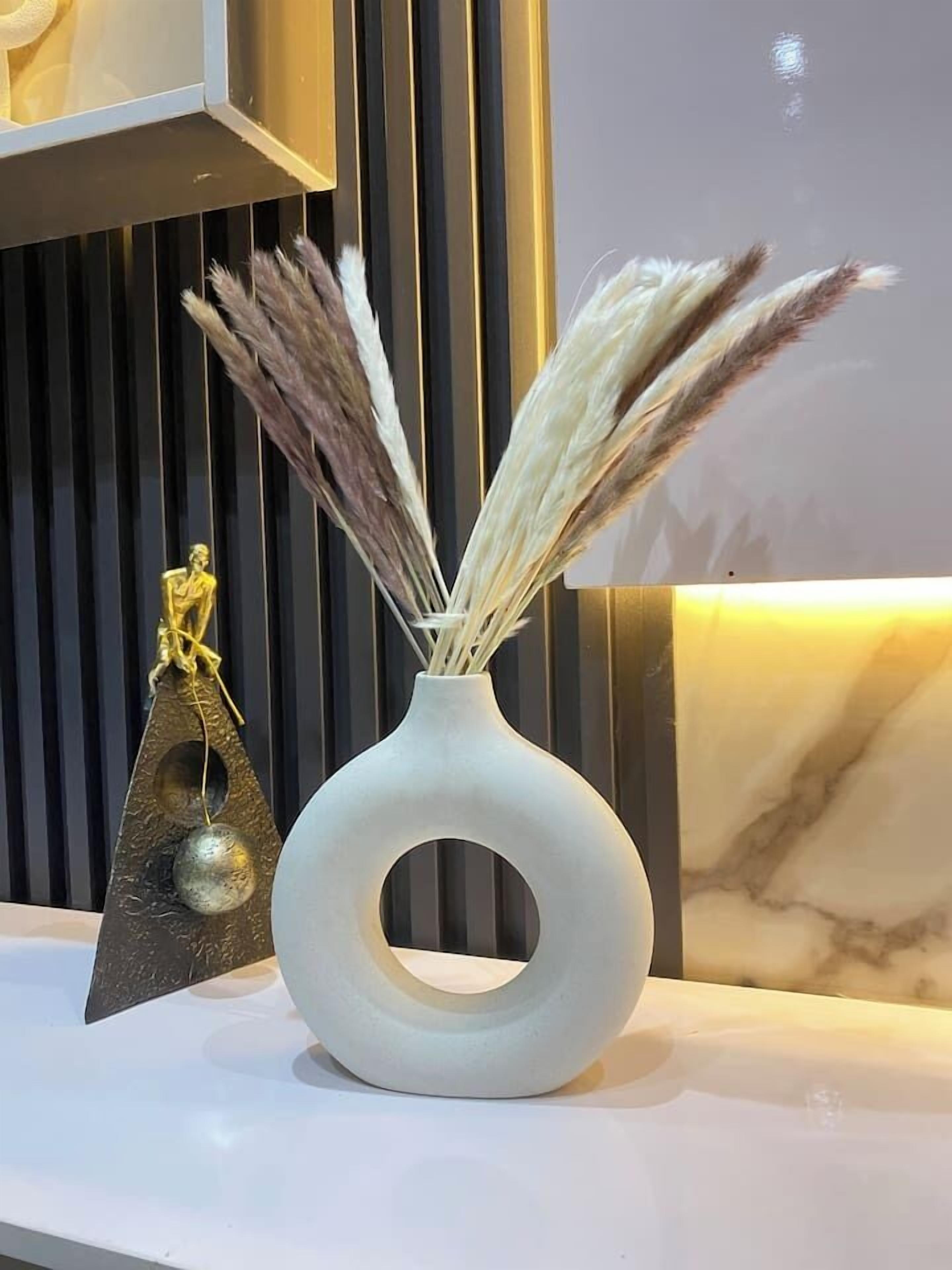 A white vase with a circular cutout containing dried grasses, placed on a white surface against a dark background with vertical lines.