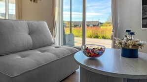 Una sala moderna con un sofá gris, una mesa redonda con un cuenco de cristal lleno de objetos coloridos y un jarrón con flores secas.