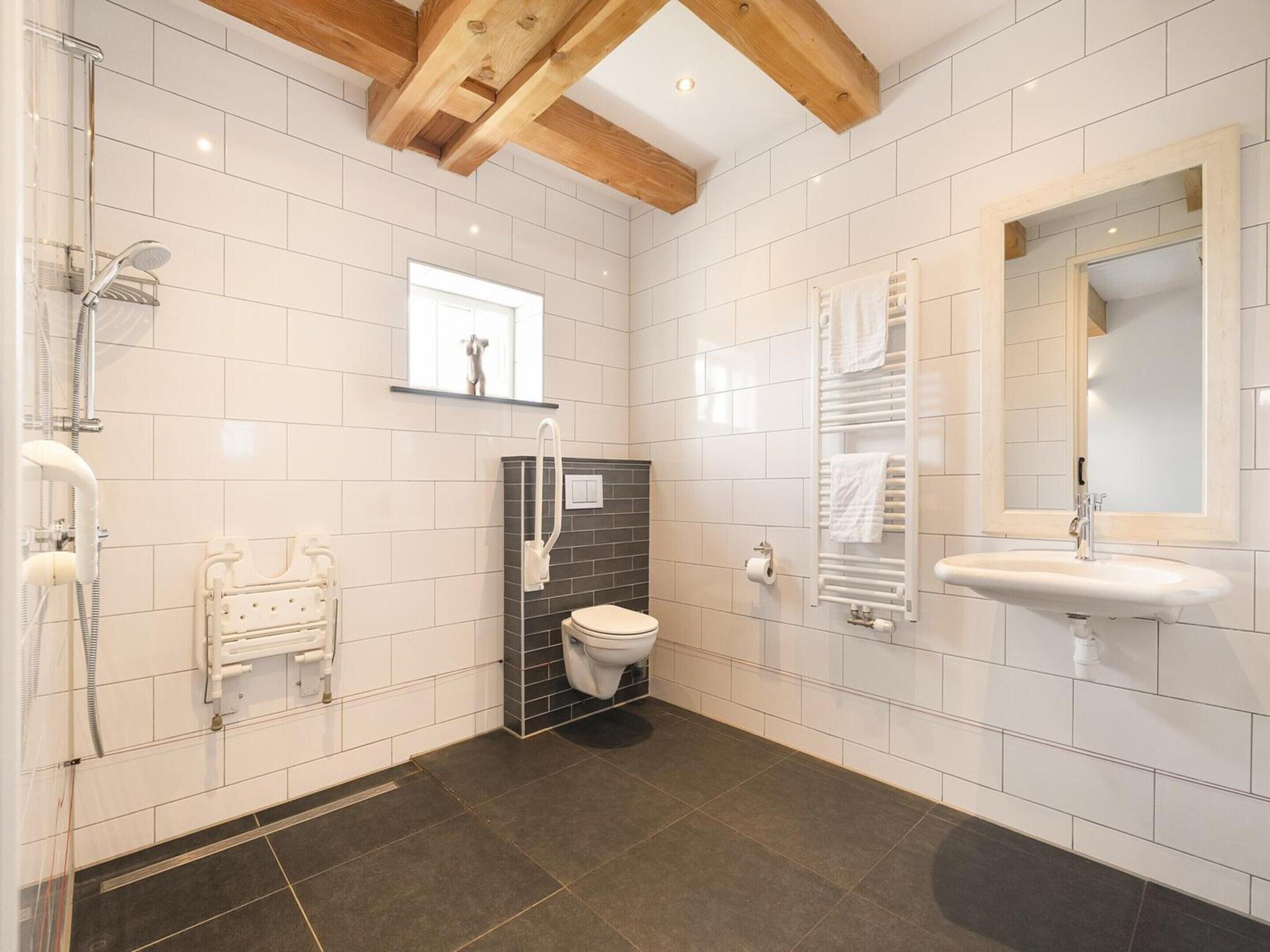 Ein modernes Badezimmer mit weißen Fliesen, einer Dusche, einer Toilette, einem Waschbecken und einem Holzbalken an der Decke.