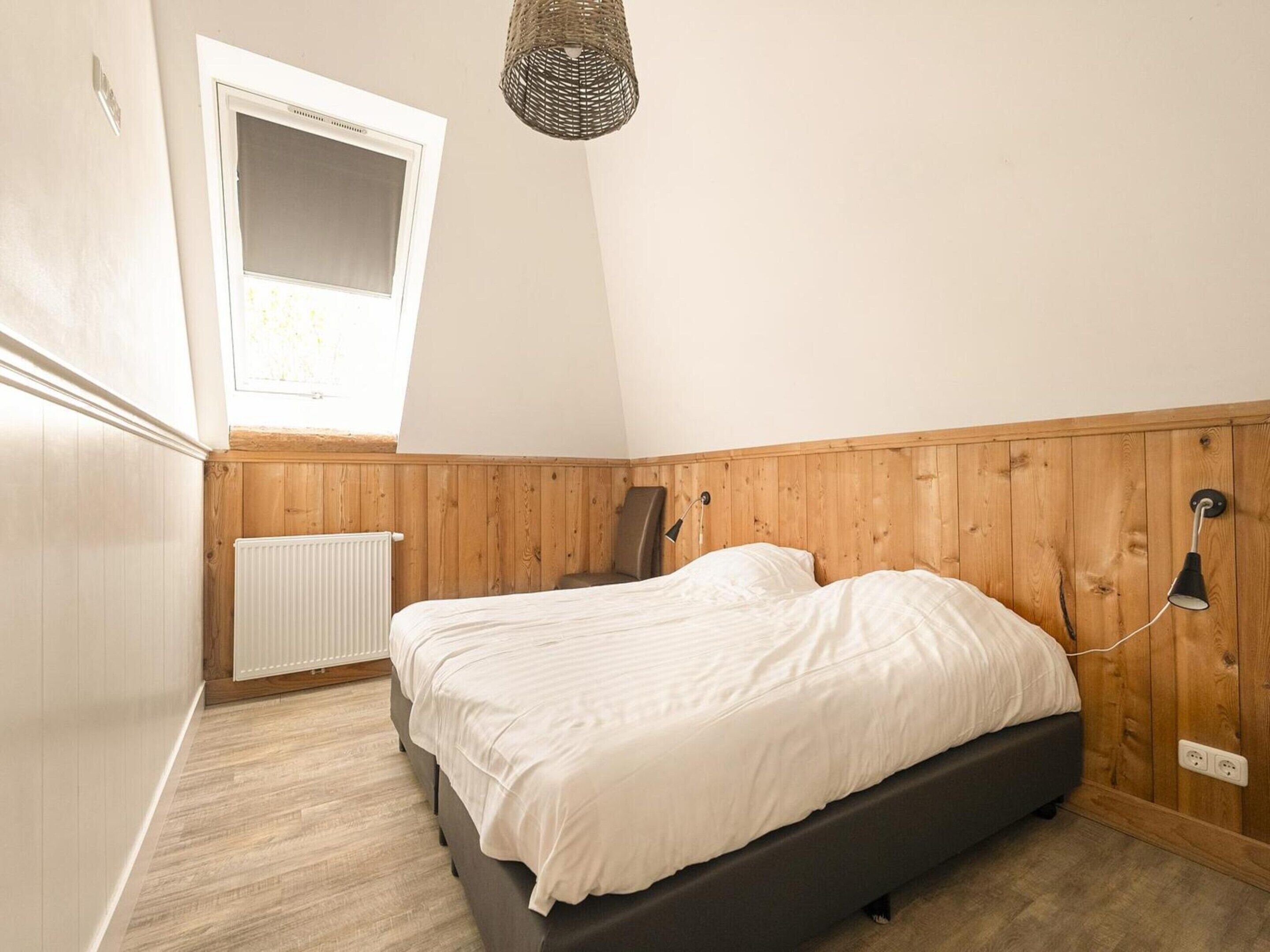 Et soveværelse med skråtloft, en seng, en radiator, et vindue og en hængende lampe.