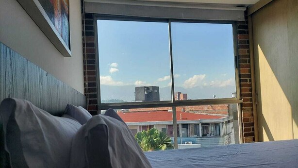 A bedroom with a large window, a bed, and a view of buildings and a red-roofed structure outside. - La 70 Laureles Condo cold AC (Medellín)