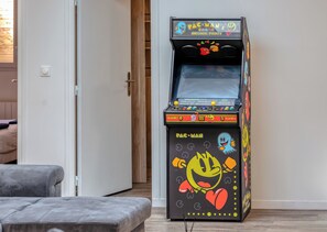 A Pac-Man arcade machine in a room with a grey couch and a wooden door. - SER LOFT 8 : Grand appartement 12 chambres idéal groupes, pros et familles (Saint-Étienne-du-Rouvray)