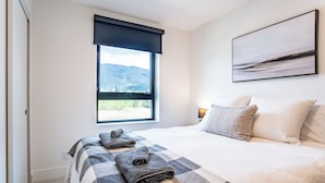 A bedroom with a large bed, a window with a view of mountains, and a painting on the wall. - Relax, Explore, Repeat –Your Revelstoke Retreat Awaits, 3 Bedroom, Pet Friendly! (Revelstoke)
