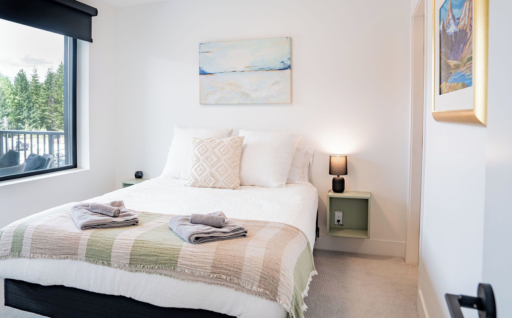 Een net opgemaakt bed met een geruit plaid, een bedlampje en een schilderij aan de muur.
