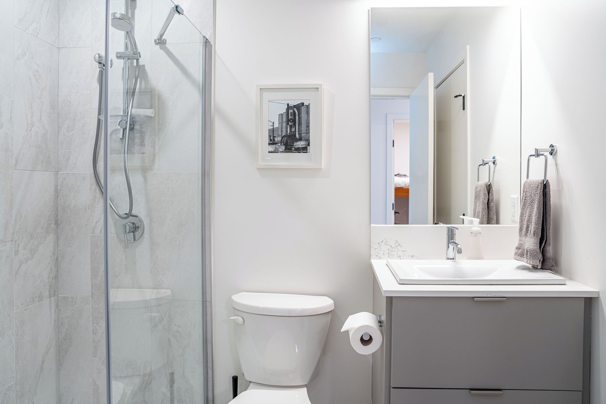 Een moderne badkamer met een glazen douchecabine, een toilet, een wastafel met spiegel en een lijst met een afbeelding aan de muur.