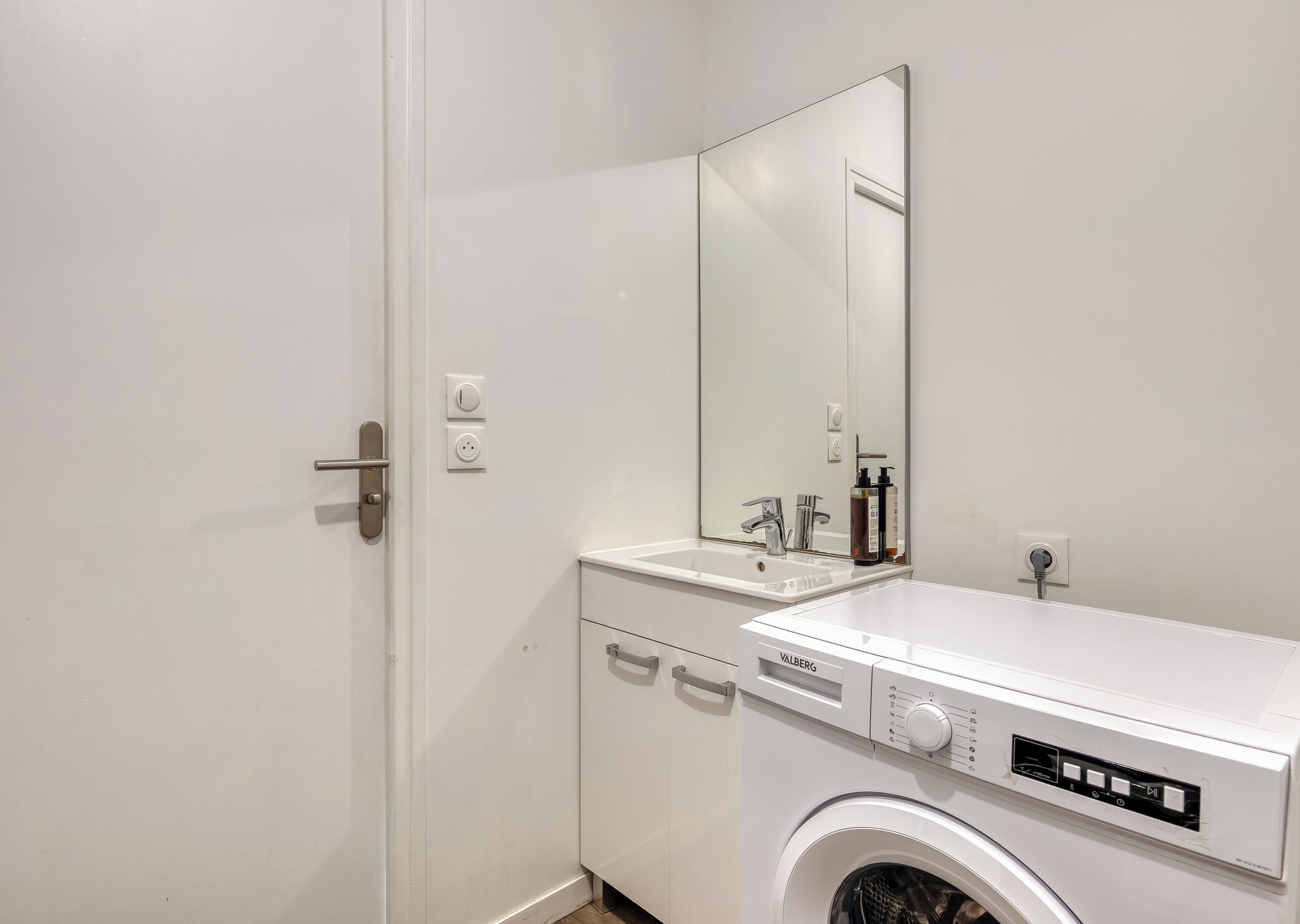 A compact bathroom with a washing machine, a sink, a mirror, and a door with a handle.