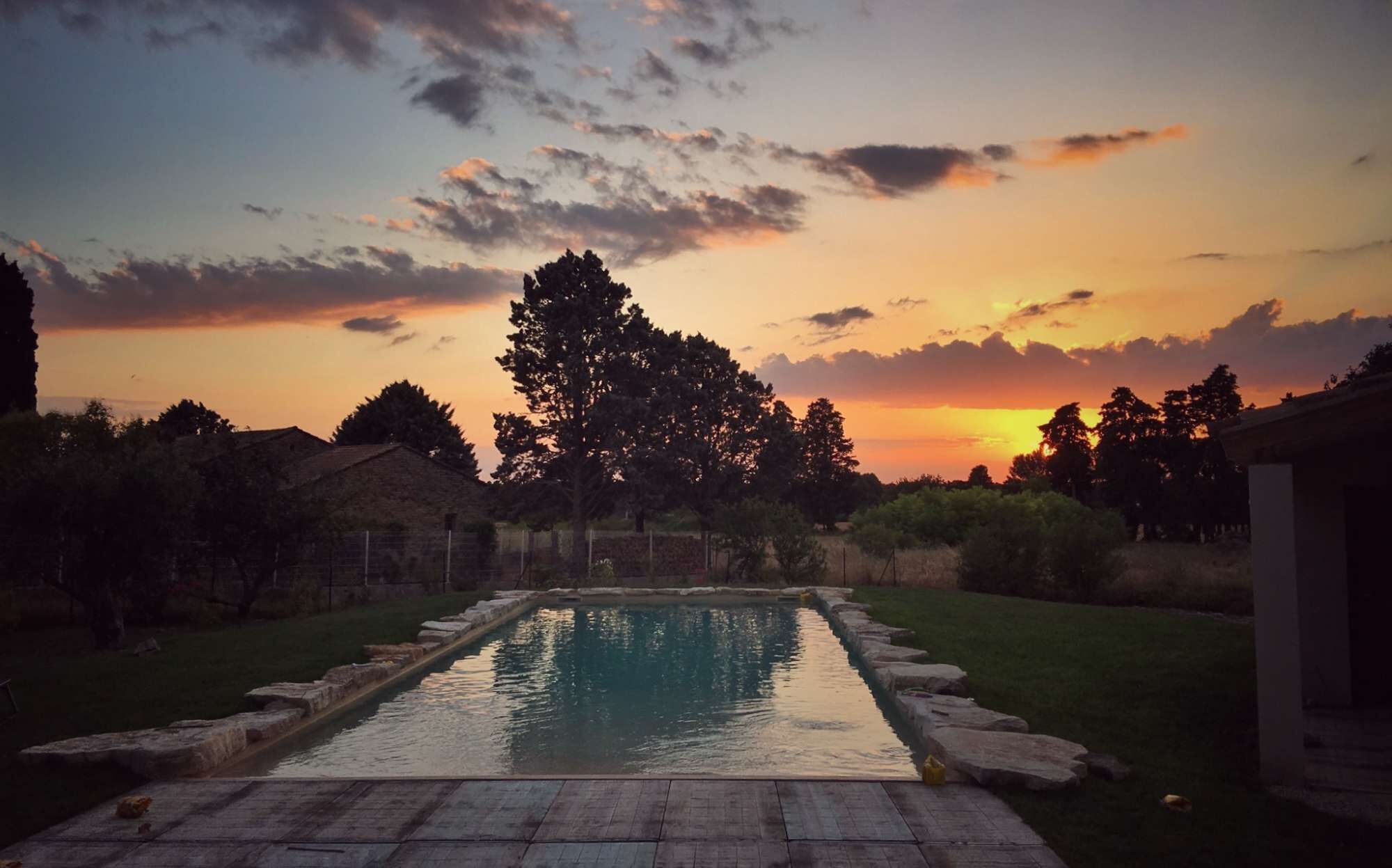 Piscina com borda de pedra, cercada por grama e árvores, sob um céu ao pôr do sol.