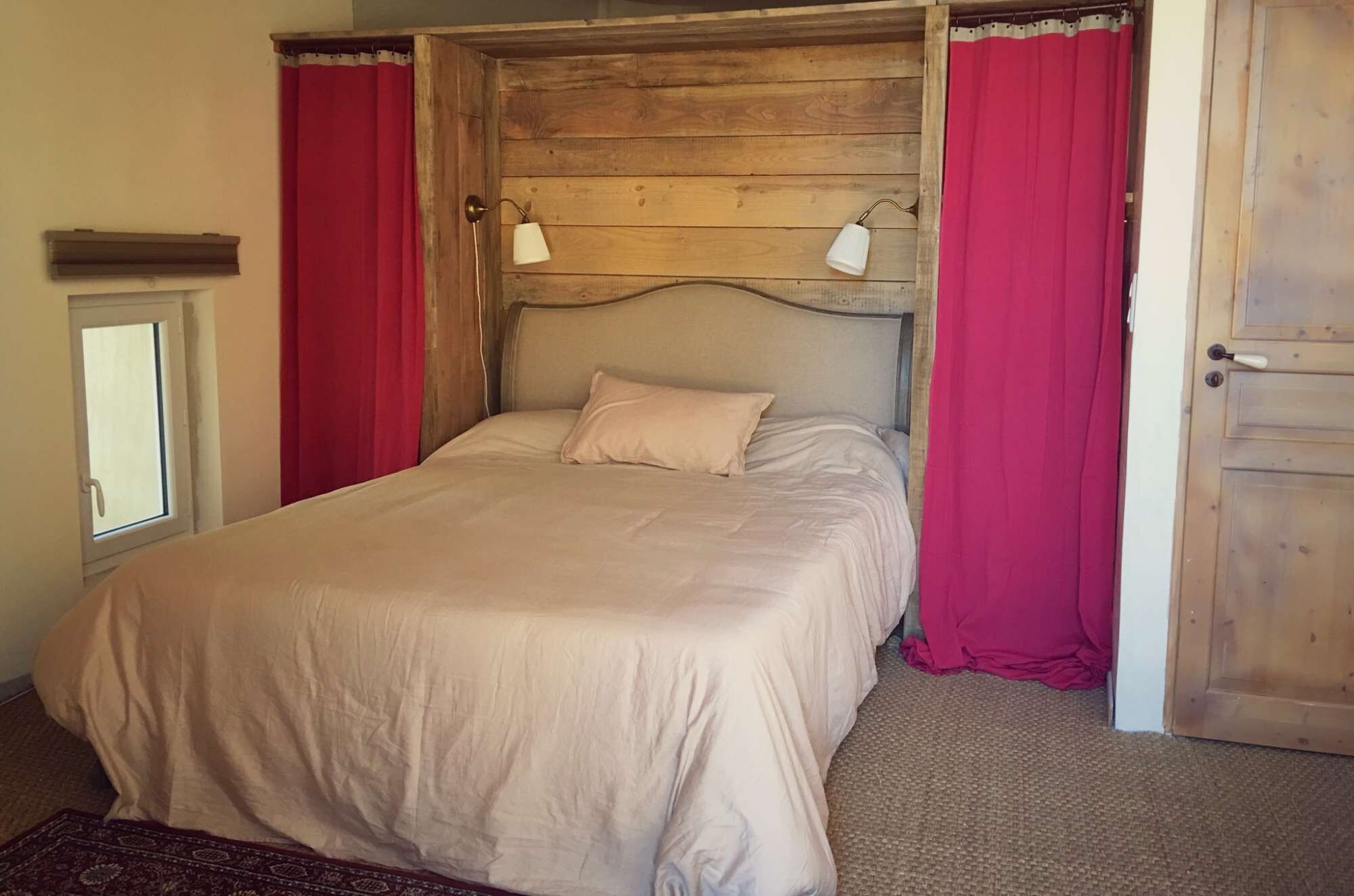 A single bed with a wooden headboard, two bedside lamps, and red curtains.