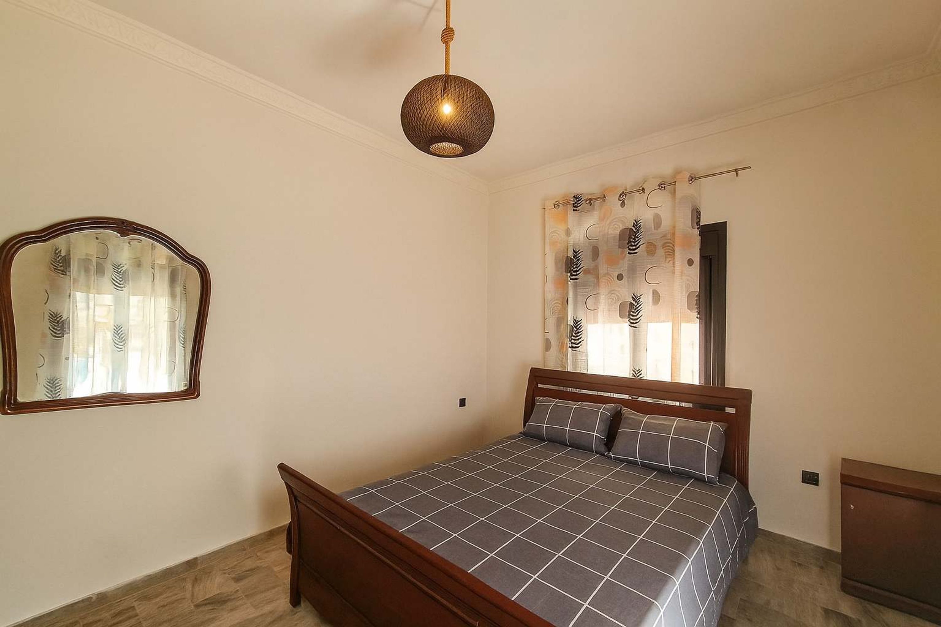 A bedroom with a wooden bed, a hanging light fixture, a mirror, and a window with patterned curtains.