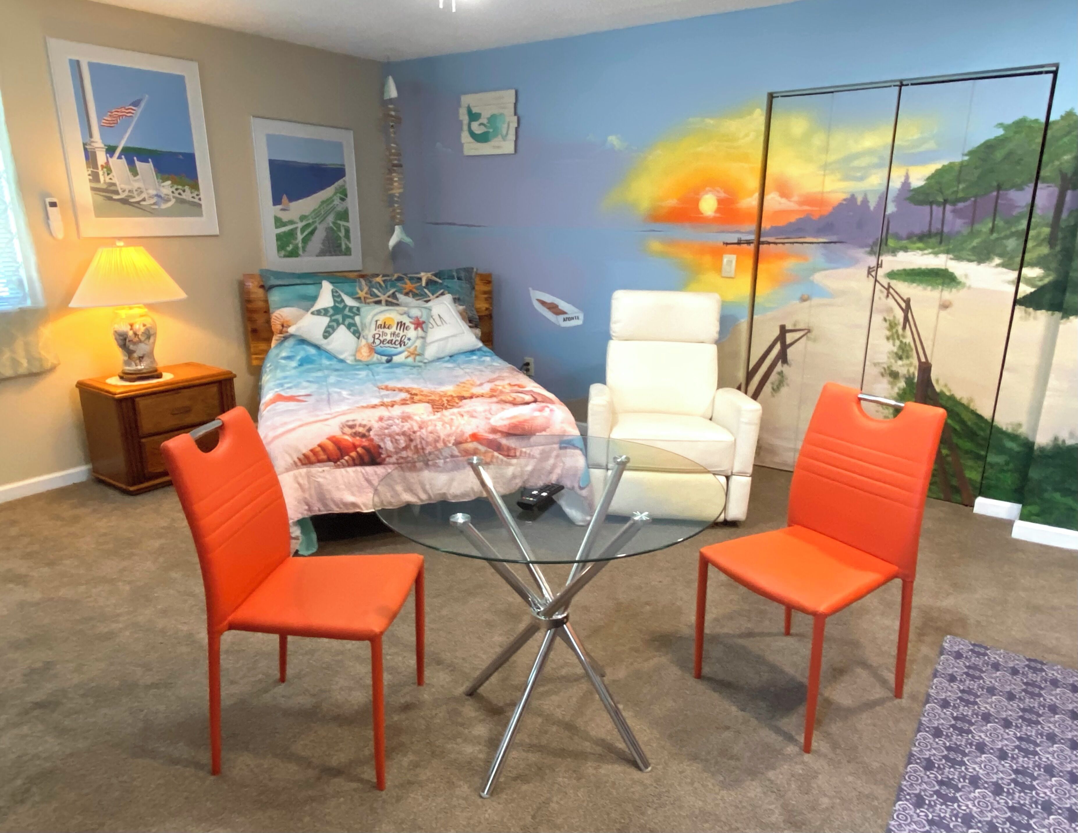 Un dormitorio con cama, dos sillas naranjas, una mesa de vidrio, una mesita de noche y una obra de arte en la pared que representa una escena de playa.