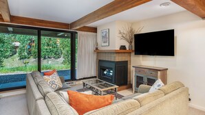 A living room with a sofa, a fireplace, a television, and a view of greenery through large windows. - Mountain Edge Condo Mt Crested Butte (Crested Butte)