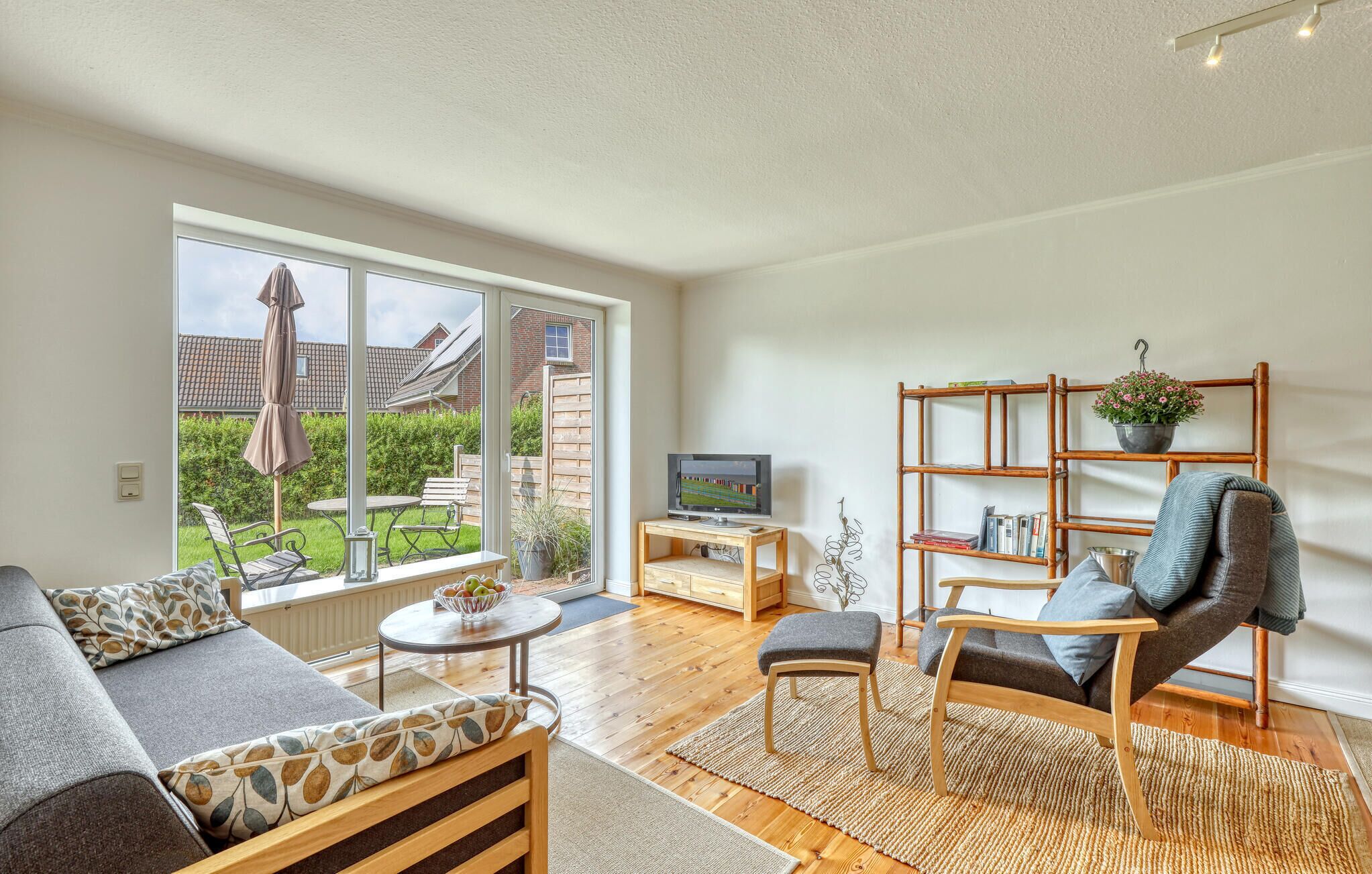 A modern living room with a sofa, armchair, and TV, large windows, and a wooden bookshelf.