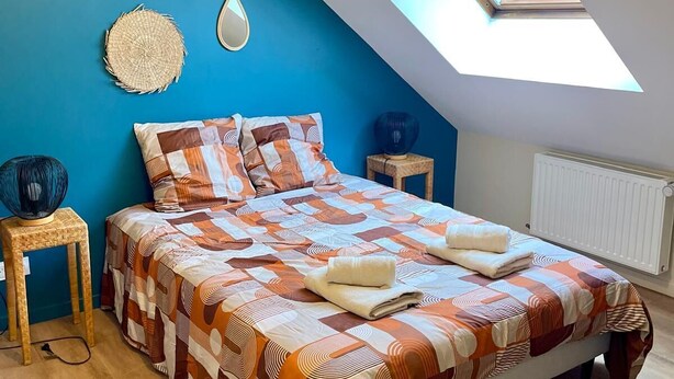 Un dormitorio con techo inclinado, tragaluz, cama con colcha estampada, mesita de noche con lámpara y espejo montado en la pared.