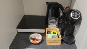 Una kitchenette compacta con encimera negra, cafetera, un tazón pequeño, un caja de madera con cápsulas de café y un refrigerador blanco.