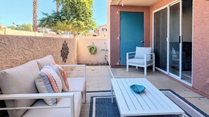 Une terrasse avec un canapé blanc, une table blanche et un tapis bleu. On y trouve une porte et une fenêtre donnant sur des palmiers et un immeuble.