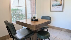 Un coin repas avec une table en bois, des chaises noires, un luminaire suspendu, une fenêtre donnant sur un espace verdoyant et un tableau encadré au mur.