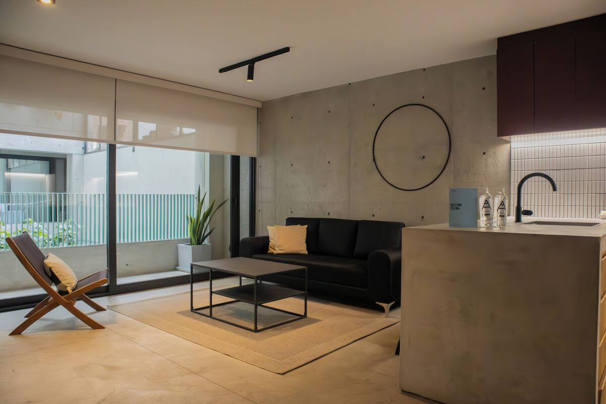 A modern living room with a sofa, a coffee table, and a chair. There is a kitchen area with a sink and cabinets.