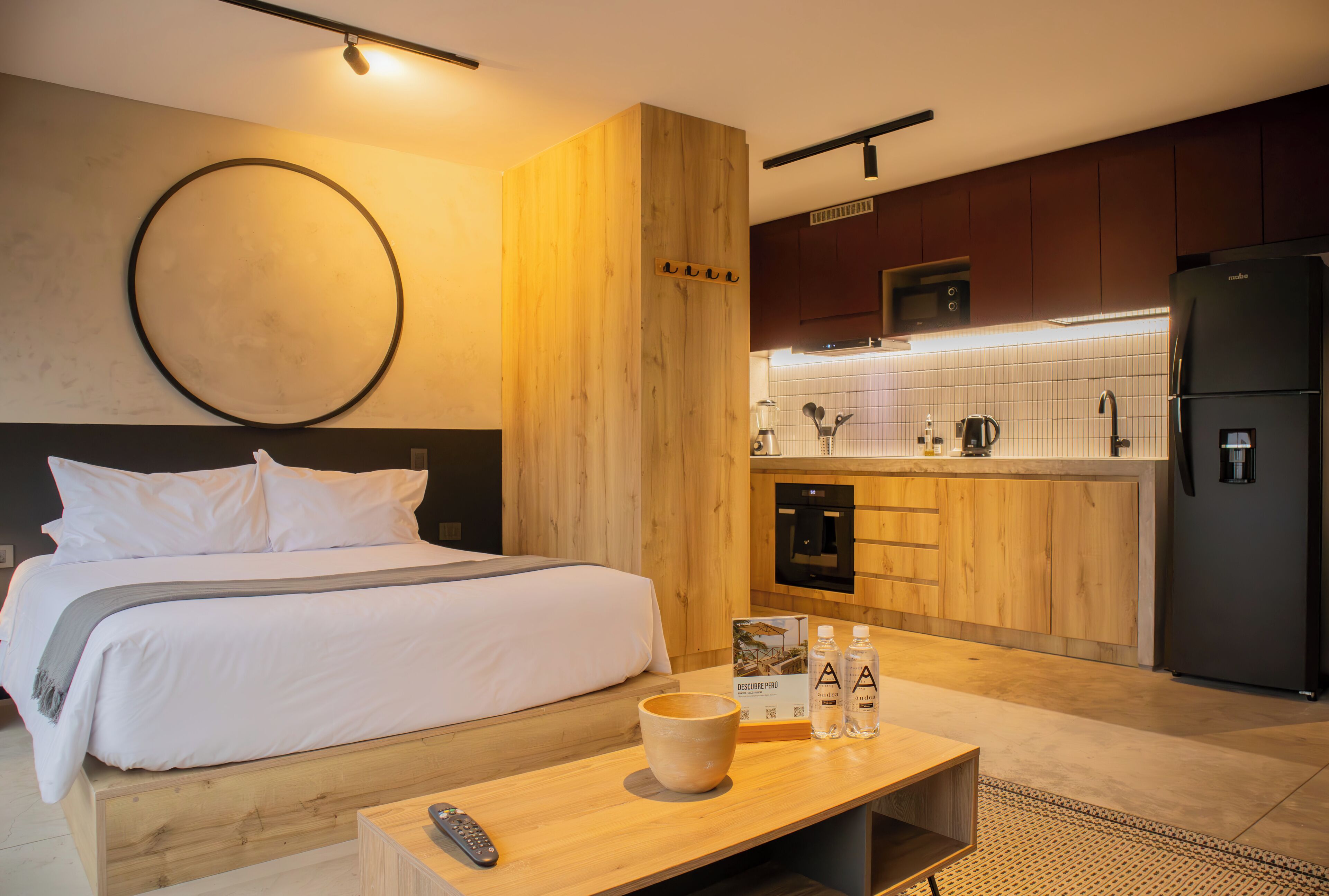 A modern hotel room with a wooden bed, a small kitchen area, and a wooden nightstand with a remote control and a bowl.