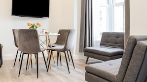 A modern living room with a flat-screen TV mounted on the wall, a round dining table with two chairs, and a grey sofa. - Baker Chambers (Camborne)