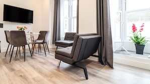 A modern living room with a flat-screen TV, grey upholstered chairs, a small table with a vase of flowers, and a large window with curtains. - Baker Chambers (Camborne)