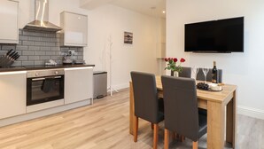 Cozinha moderna com forno embutido, exaustor de aço inoxidável e painel de azulejos. Há uma área de refeições com mesa e cadeiras de madeira, e uma TV de tela plana na parede.