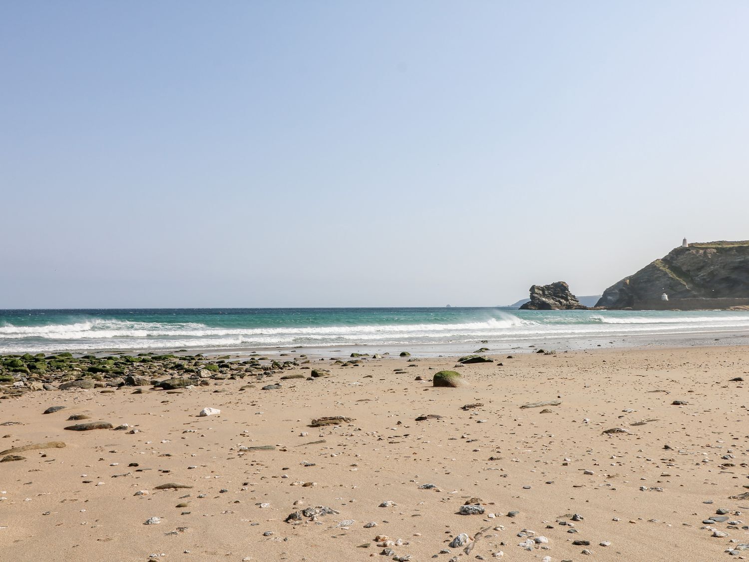 A beach with scattered rocks and a lighthouse on a cliff in the background.