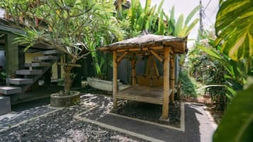 A wooden gazebo with a thatched roof, surrounded by tropical plants and a gravel path.