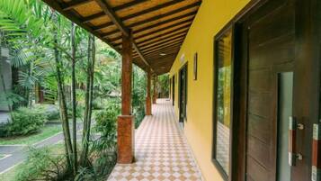 A covered walkway with a checkered floor, surrounded by greenery and a building with a dark door.